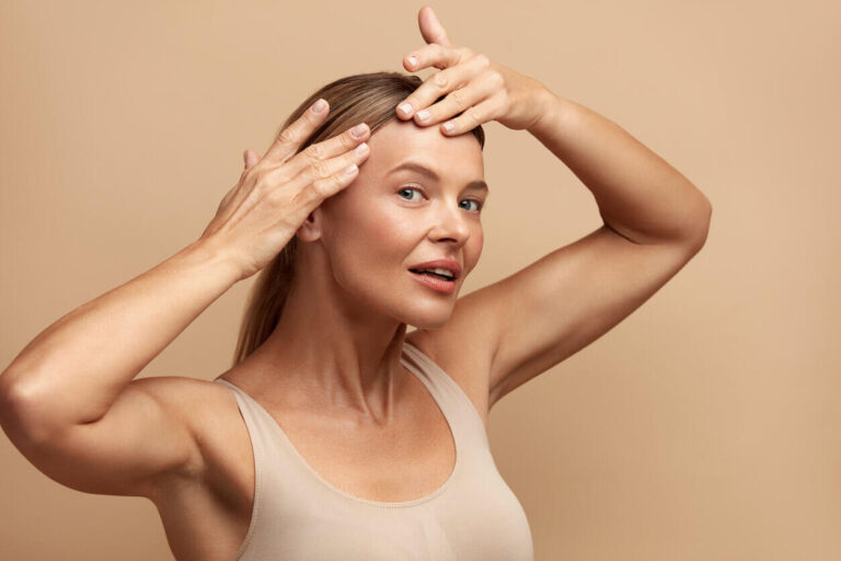 Mulher madura tocando a testa com as mãos e sorrindo levemente, representando cuidados preventivos contra rugas e envelhecimento da pele