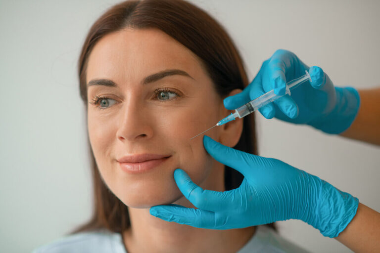 Mulher sorrindo enquanto recebe uma aplicação de botox preventivo na região do rosto, realizada por profissional com luvas azuis em ambiente clínico.
