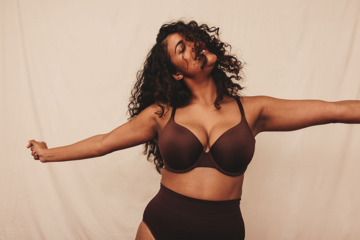 Mulher de cabelos cacheados usando lingerie marrom, com expressão de liberdade e braços abertos, simbolizando autoestima e bem-estar após cirurgia estética — representando os benefícios da mastopexia na confiança e na valorização do corpo feminino.