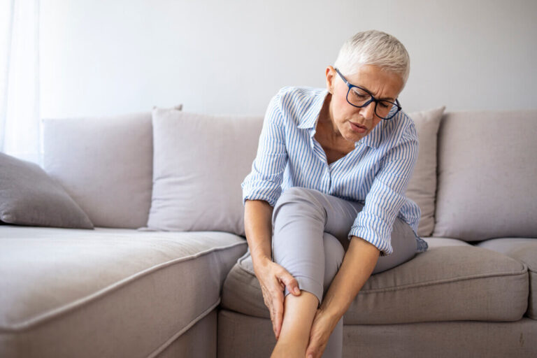 Mulher madura sentada no sofá segurando a perna com expressão de dor, representando os desconfortos que podem surgir com a menopausa e lipedema.