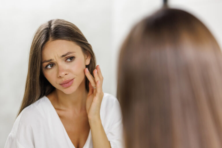 Mulher observando o rosto no espelho, representando avaliação estética após excesso de preenchimento facial.