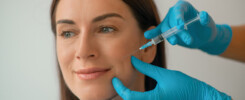 Mulher sorrindo enquanto recebe uma aplicação de botox preventivo na região do rosto, realizada por profissional com luvas azuis em ambiente clínico.