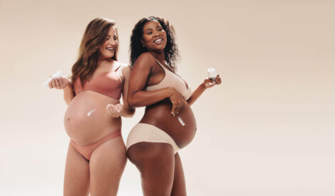 Duas mulheres grávidas sorriem enquanto aplicam creme na barriga, representando casos de gestação saudável após uma abdominoplastia pós-bariátrica, mostrando confiança e bem-estar corporal.